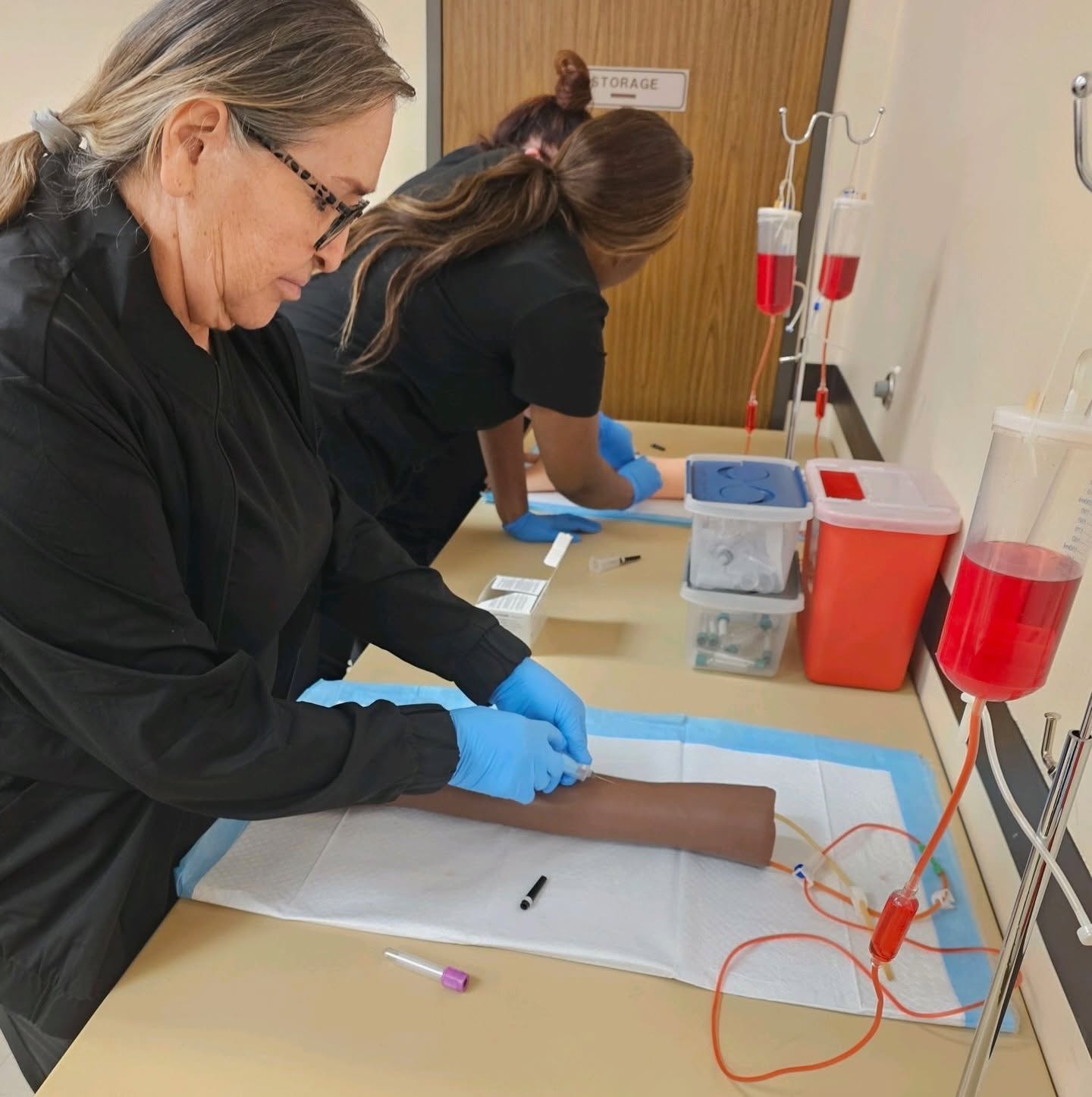 Two students in black scrubs practicing IV insertion on a training arm with IV pole and sharps container