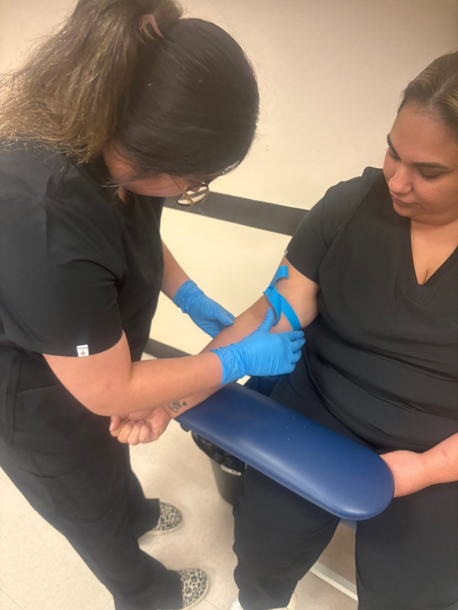 Student in blue gloves practicing tourniquet placement and venipuncture technique on a fellow student