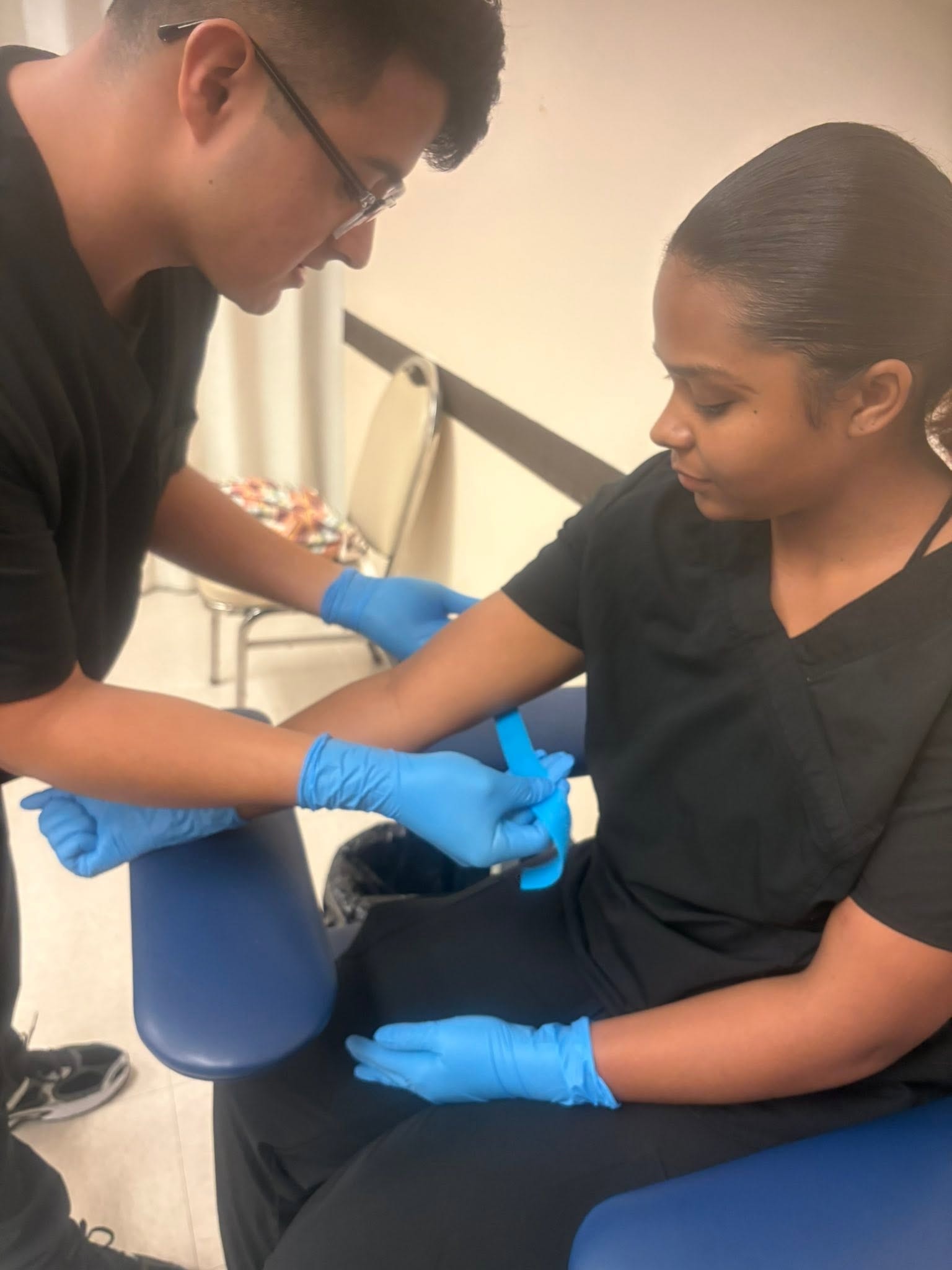 Student in blue gloves applying a tourniquet and preparing for a venipuncture draw on a fellow student